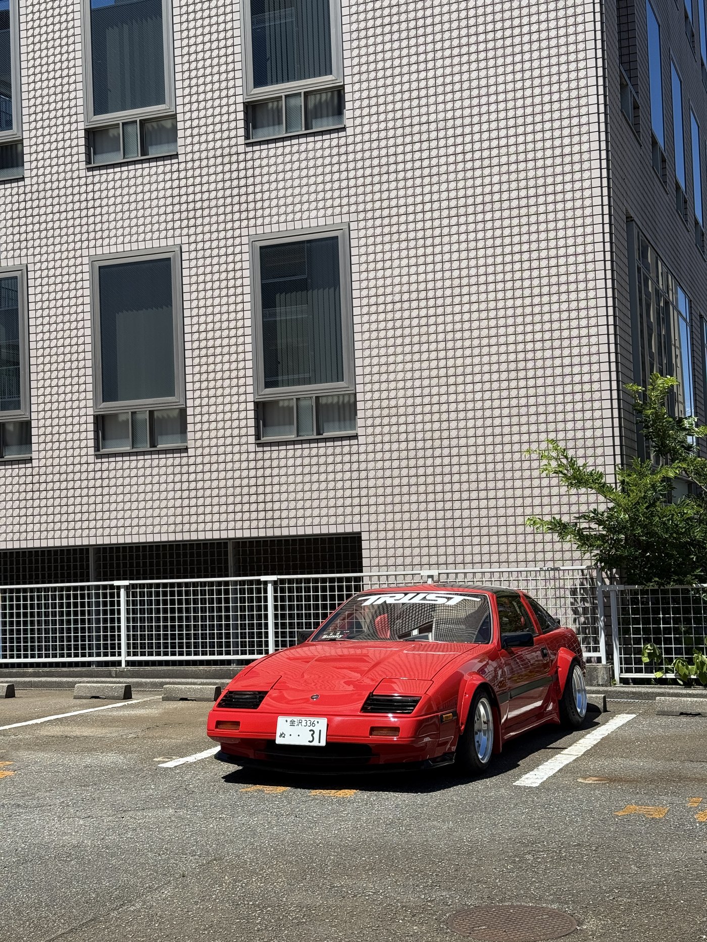 Červená Mazda RX-7 se samolepkou TRUST před obloženou budovou